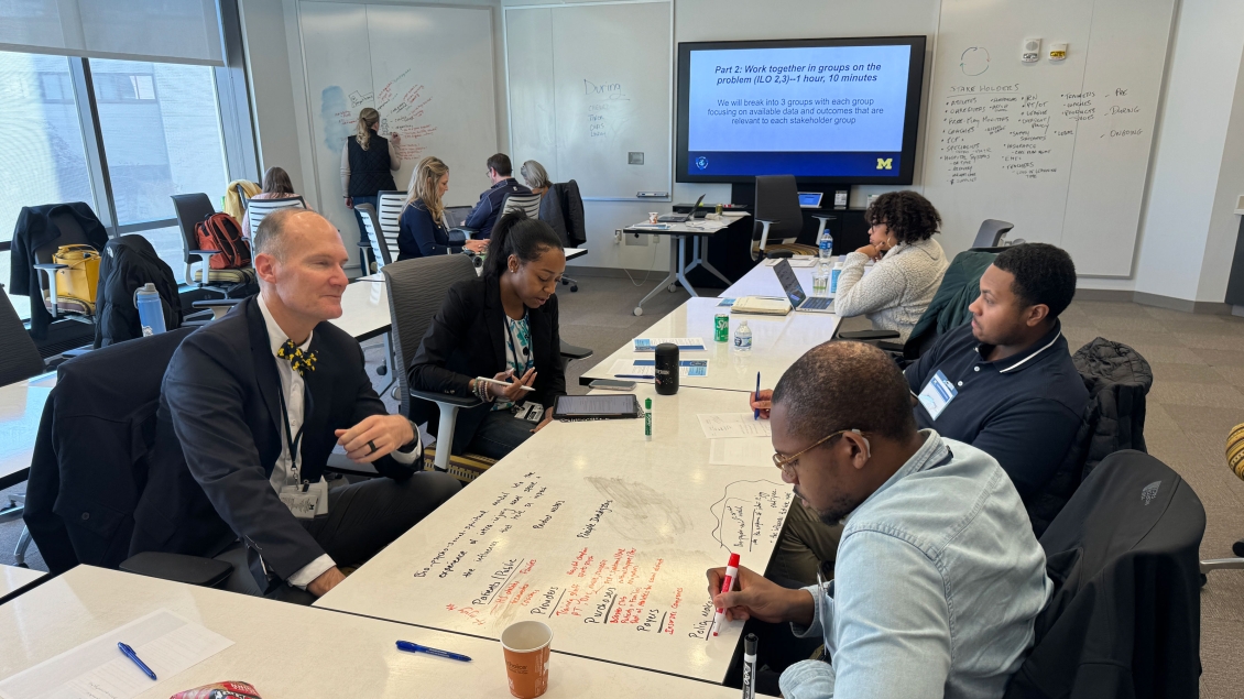 A collaborative group discussion in a professional setting, with participants actively engaging in problem-solving at a whiteboard and taking notes at a table. 
