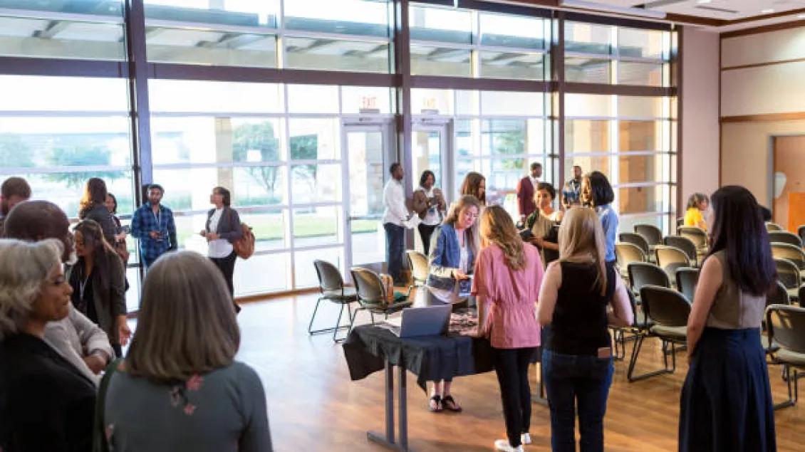 A diverse group of people mingles in a sunlit conference room.