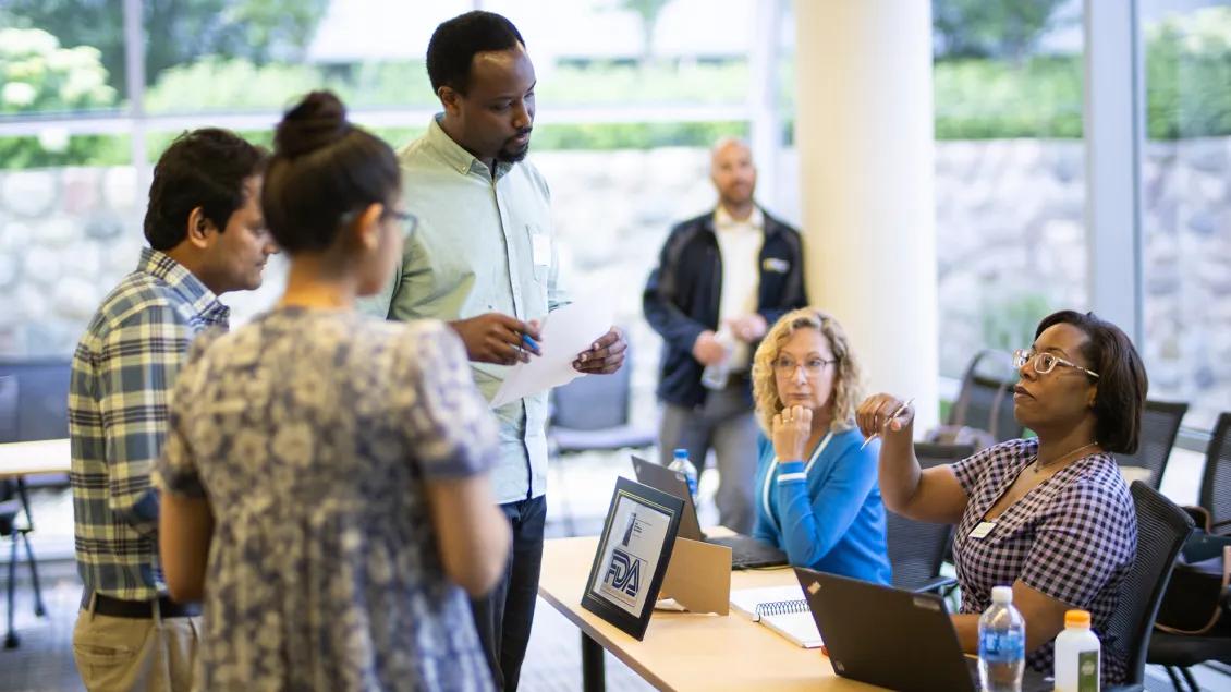 A diverse group of adults engages in a discussion around a table with laptops.