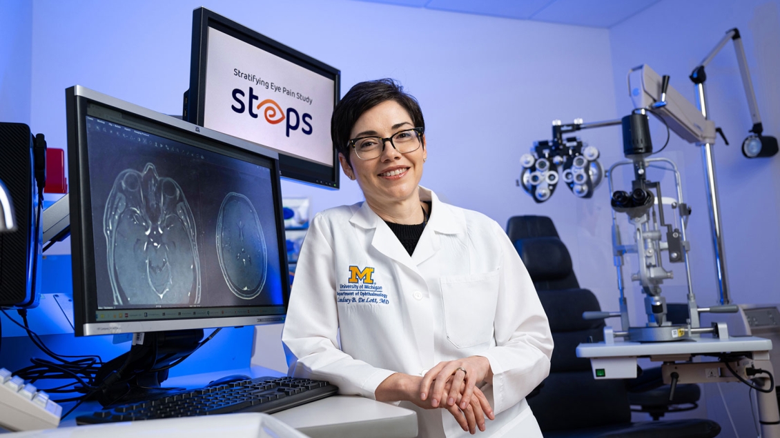 A female medical professional wearing a white University of Michigan lab coat stands smiling in a clinical setting. She is surrounded by ophthalmology equipment, including a slit lamp and phoropter, and two computer monitors—one displaying brain scans and the other showing the logo for the “Stratifying Eye Pain Study (STEPS).