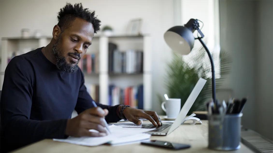 man writing at desk with laptop and paperwork