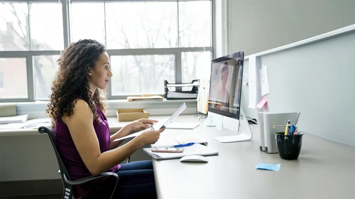 woman sitting at desk holding papers and listening to a remote meeting