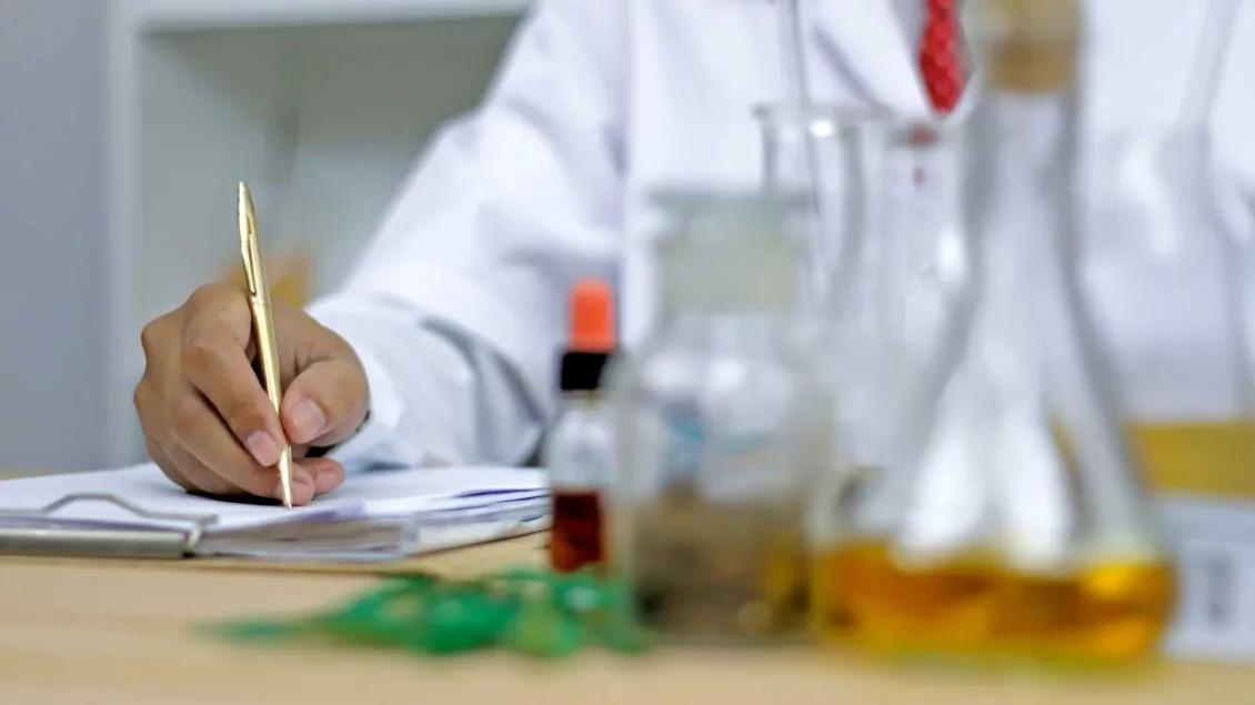 Scientist in white ccoat writing in lab journal with beakers on table