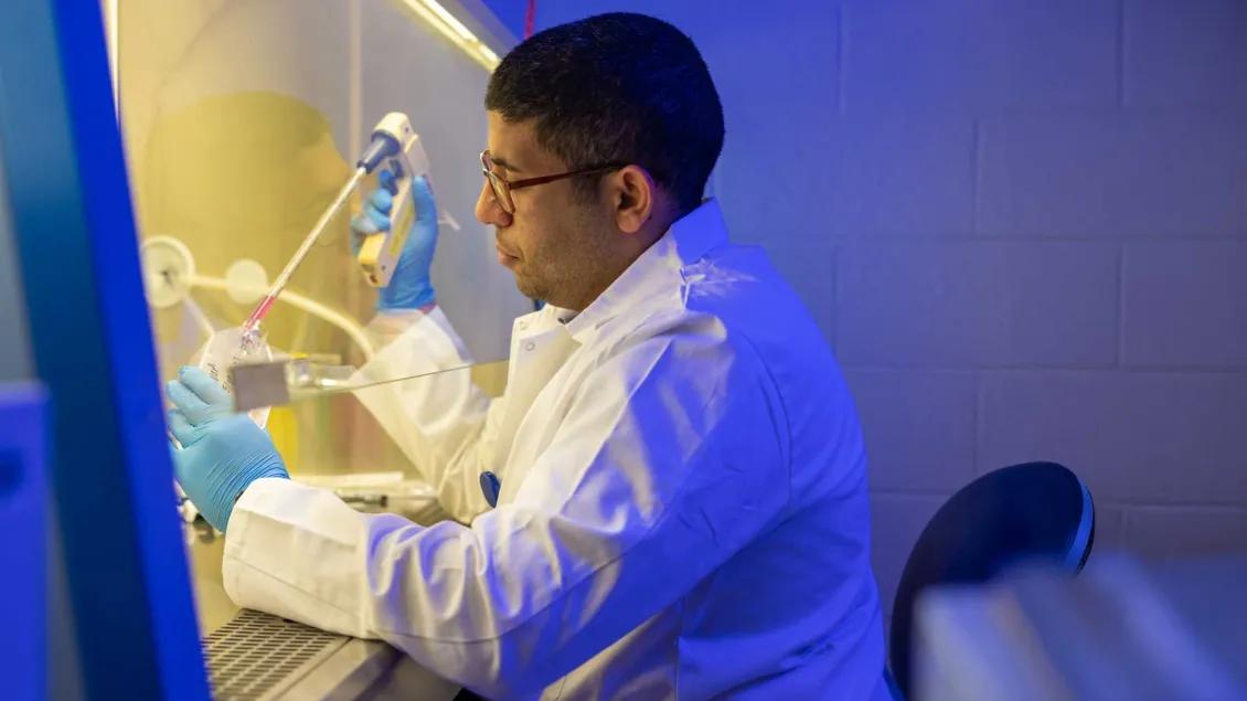 A scientist wearing a white lab coat, blue gloves, and glasses works in a laboratory biosafety cabinet, using a pipette to transfer liquid into a labeled plastic container under sterile conditions. Blue and yellow lighting highlights the controlled environment.