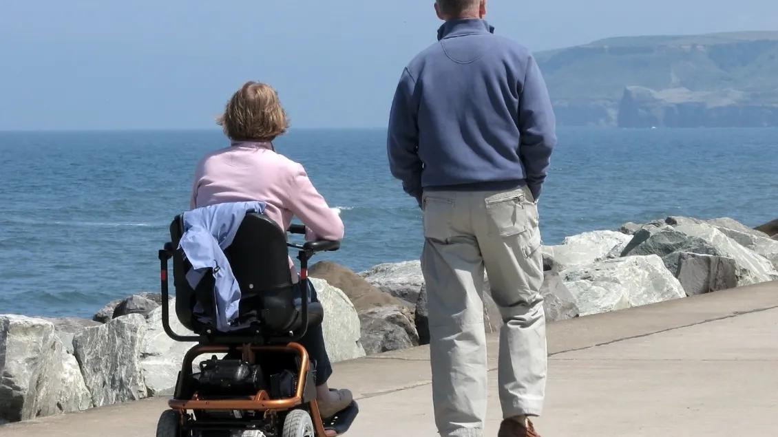 Woman in wheelchair and man walking at the sea