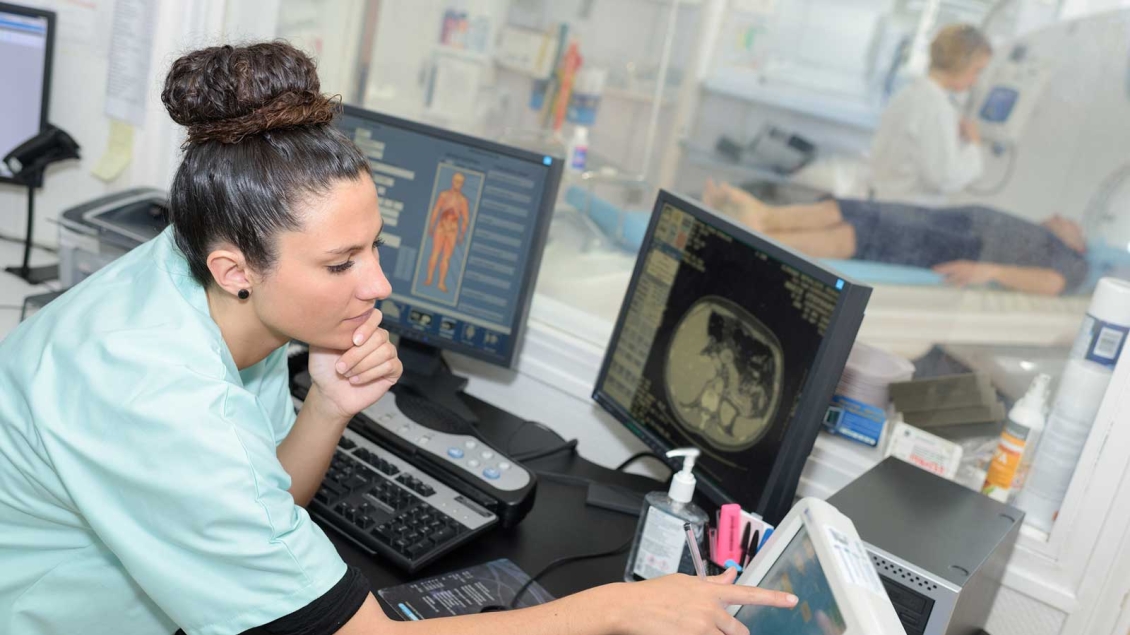 Technician reviews medical scans on monitors while a patient undergoes imaging.