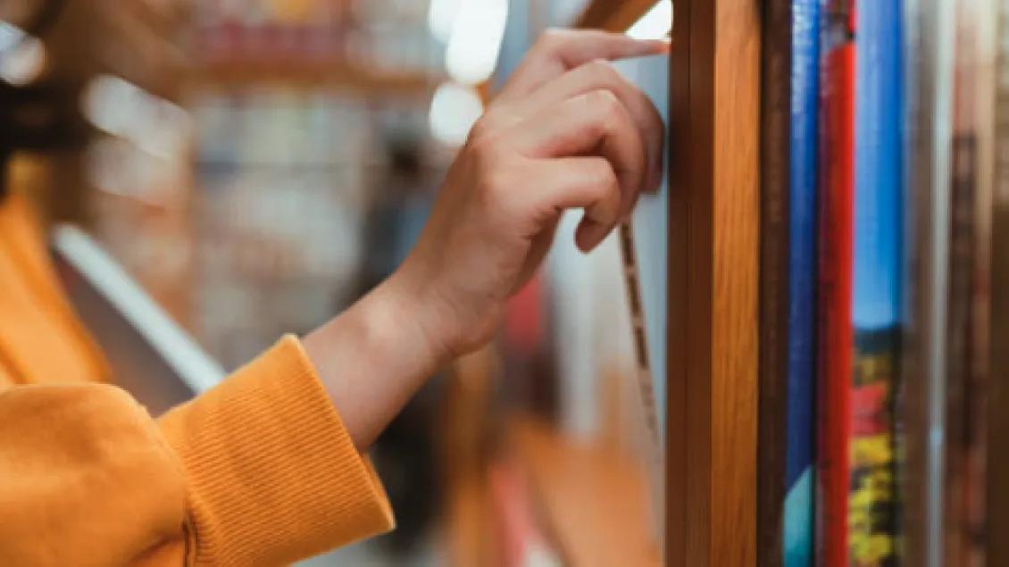 A person in a yellow sweater selects a book from a wooden shelf in a library.