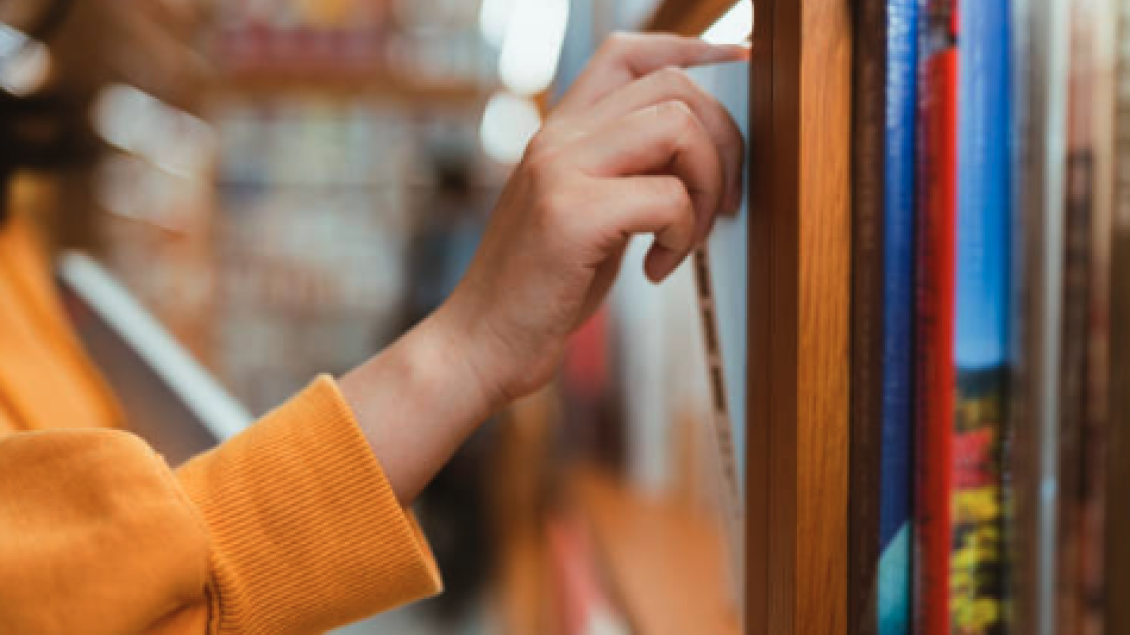 A person in a yellow sweater selects a book from a wooden shelf in a library.