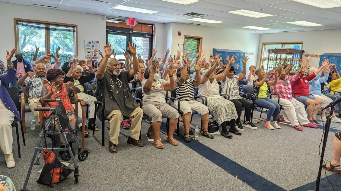A large group of older adults seated in a community room, smiling and raising their arms together in an exercise or group activity. Some people use walkers or mobility aids. The atmosphere appears lively and engaged. The room has large windows, fluorescent lighting, and blue chairs arranged in rows.