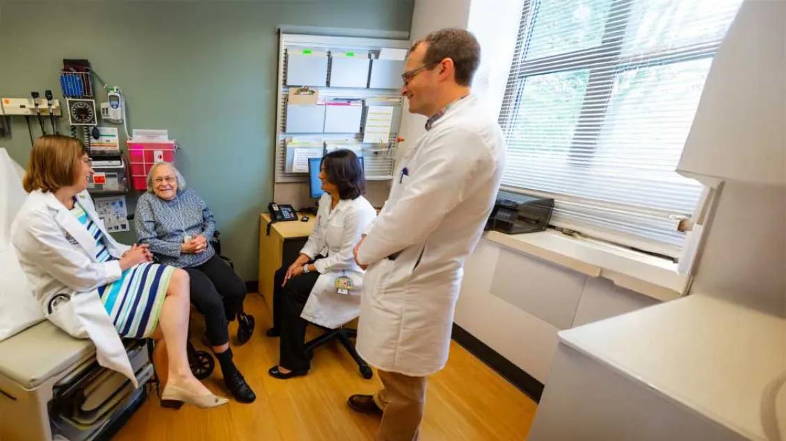 A group of healthcare professionals, including two female and one male doctor, are gathered in a medical exam room with an elderly woman sitting in a wheelchair. One doctor sits on the exam table engaging warmly with the patient, while the others listen attentively and smile, creating a collaborative and supportive care environment.