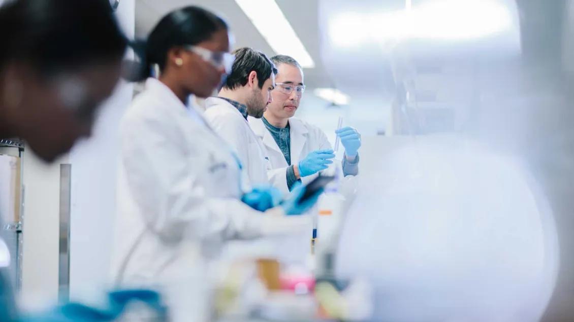 Researchers in white lab coats and blue gloves work together in a laboratory. One person holds a test tube while others examine data on a tablet. The lab is brightly lit and filled with scientific equipment.
