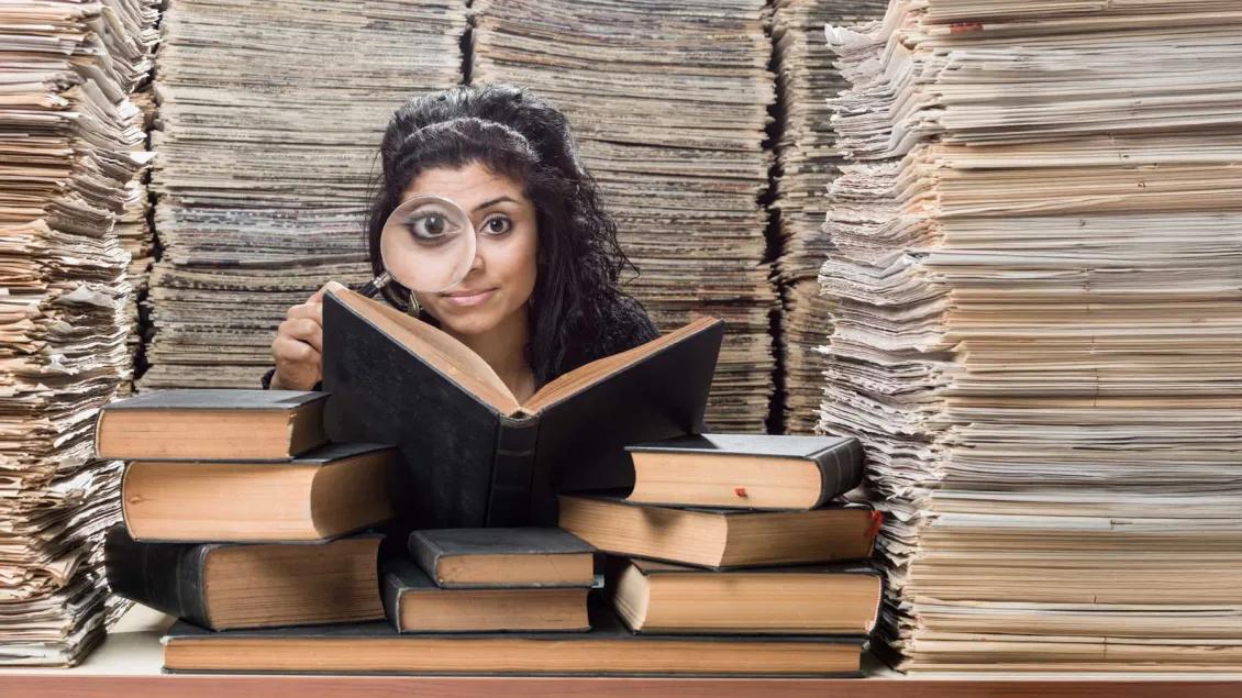 Person with a magnifier looking outward. An open book is in front of her with multiple books on a table and piles of paper all around her.