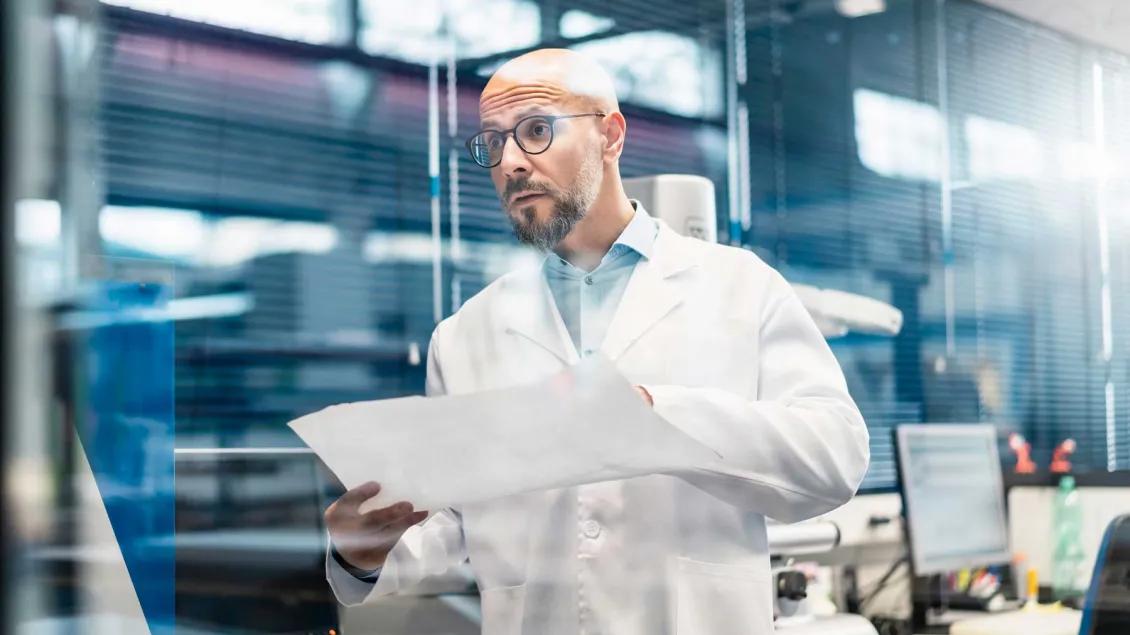 A scientist in a white lab coat and glasses examines a large data document in a modern laboratory with computers and scientific equipment in the background.