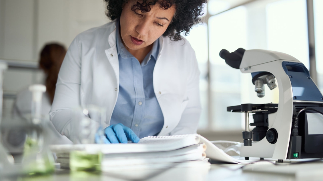 A scientist in a white lab coat is writing notes in a laboratory notebook while seated at a lab bench. A microscope is positioned beside her, and various glass containers with green liquid are visible in the foreground. She appears focused and engaged in documenting research observations.