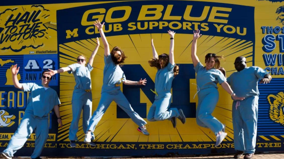 Pulmonology & Critical Care fellows jumping in front of a sign that says "Go Blue Club Supports You"