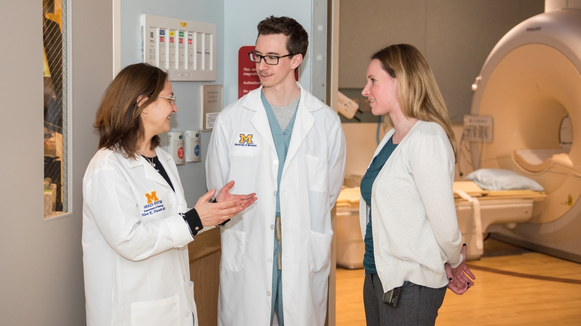 Two women and a man in white coats talk in a scan room