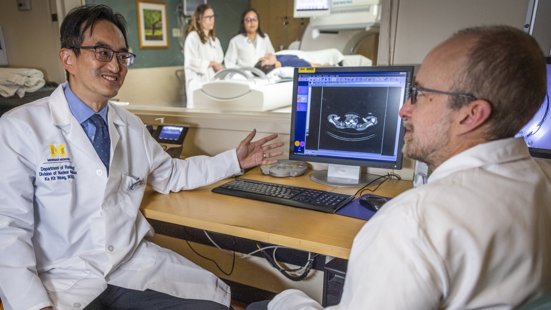 Two men in white coats talk in front of a medical scan image