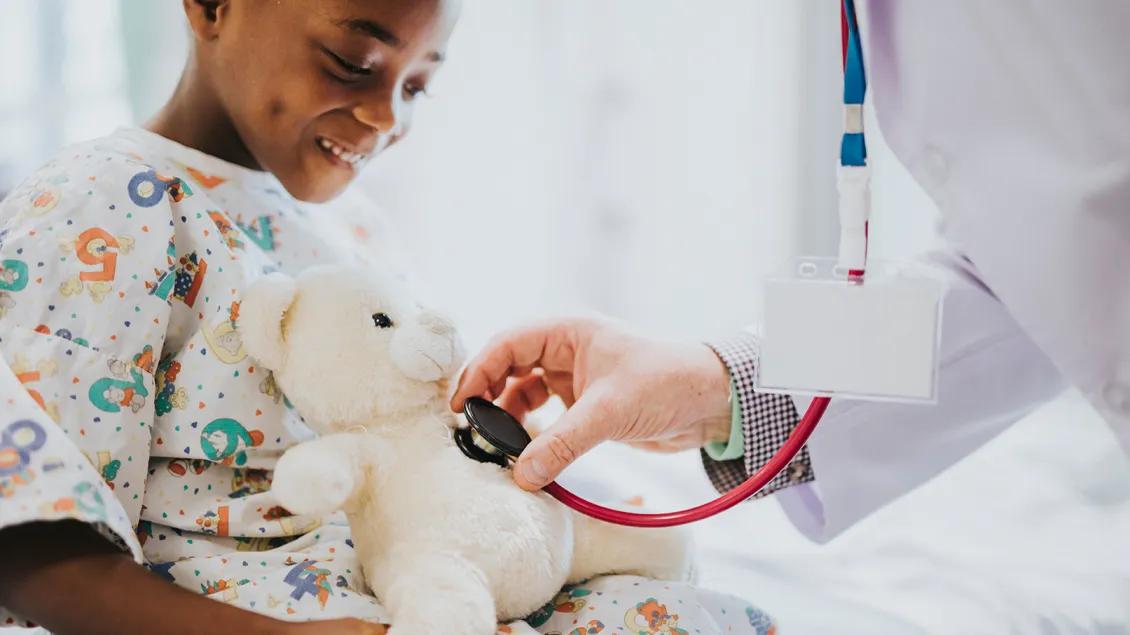 Doctor places stethoscope on teddy bear held by pediatric patient.