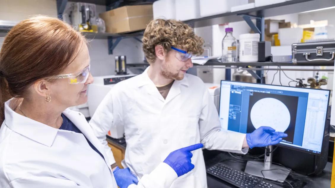 Two scientists in lab coats and safety goggles discuss data on a computer screen displaying a circular graph.