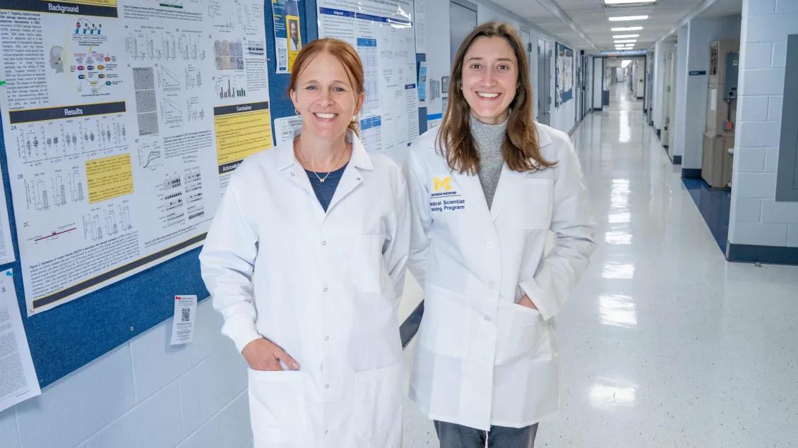Two women in lab coats stand in a bright hallway with scientific posters on the walls.