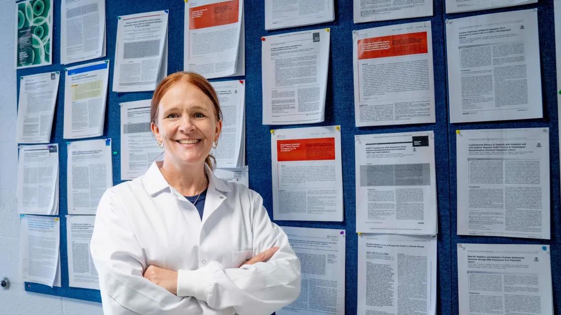 A person with red hair, wearing a white lab coat, stands confidently before a bulletin board displaying numerous papers.