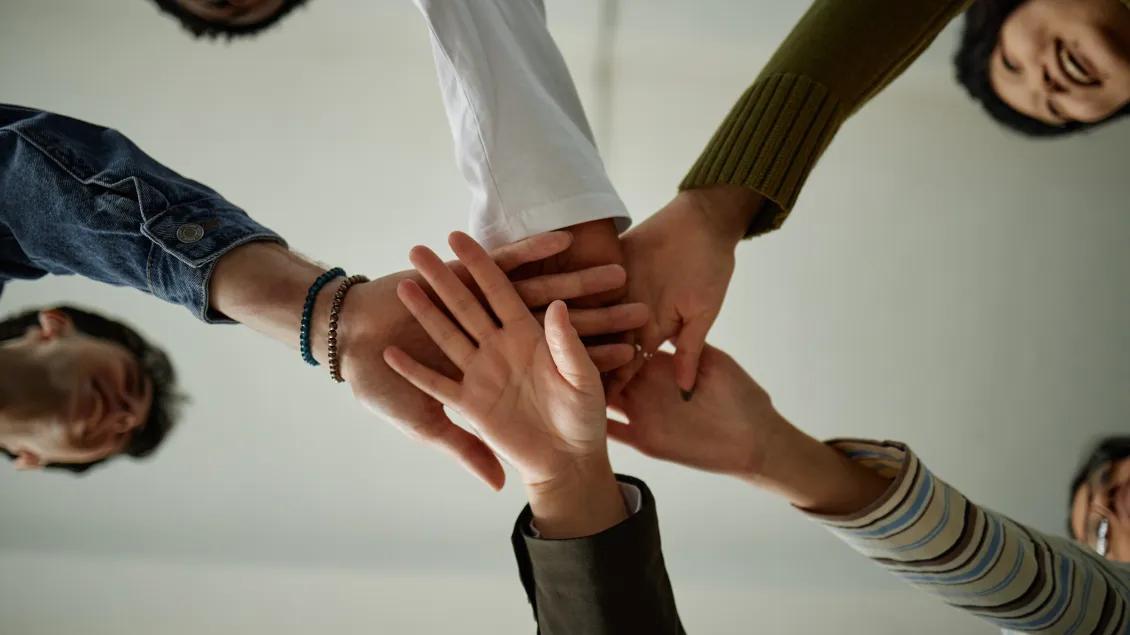 A group of people standing in a circle stack their hands together in the center, as seen from below. The individuals are wearing different clothing, such as a green sweater, a striped sleeve, denim, and white shirt, symbolizing teamwork and unity.