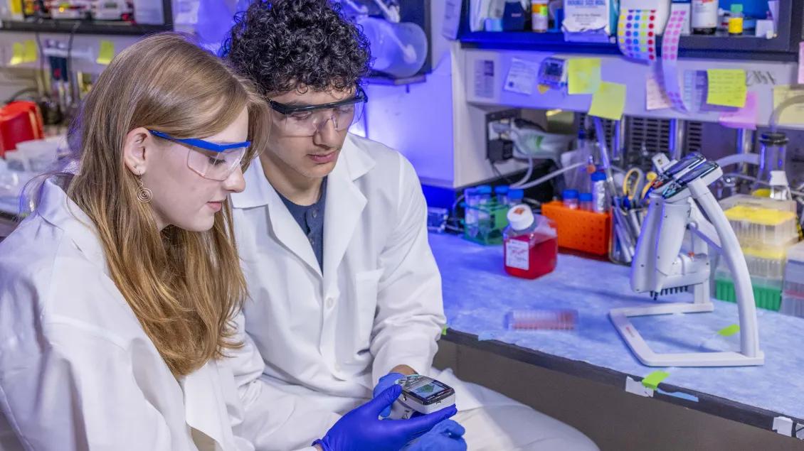 Two scientists in white lab coats and safety goggles work in a laboratory.