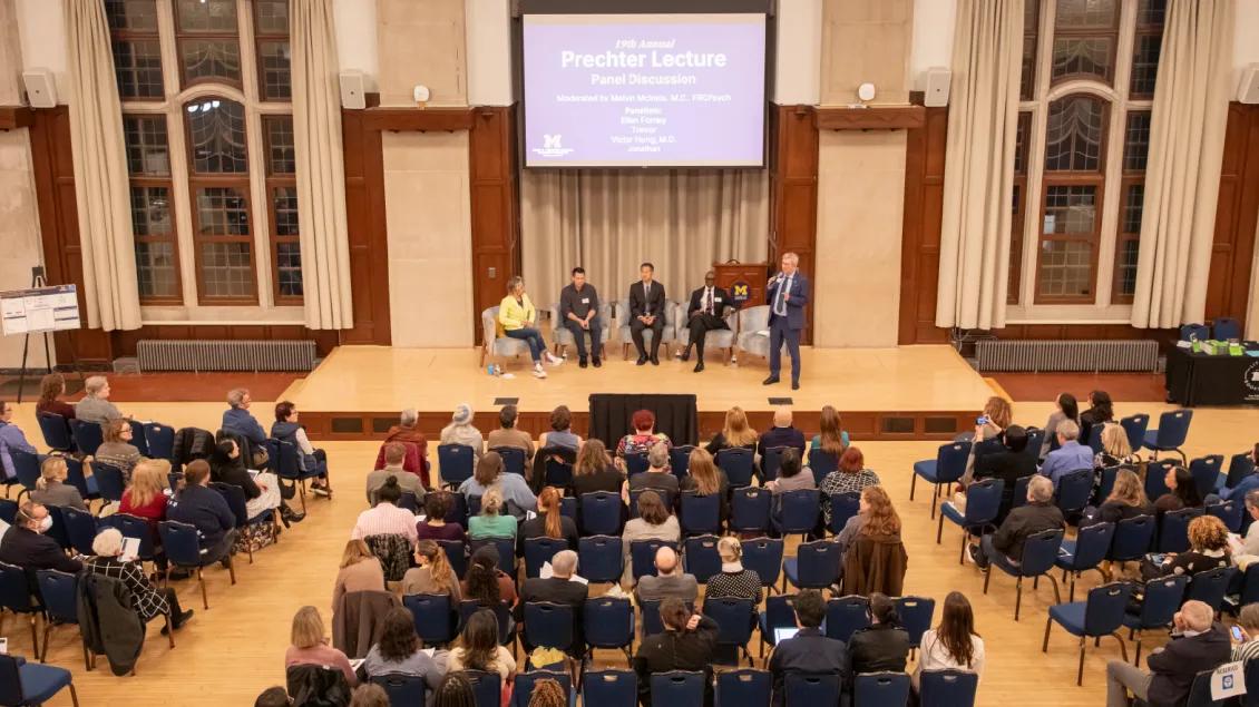A large audience sits in rows of blue chairs in a wood-paneled auditorium, facing a stage where five panelists are seated and one person is standing and speaking. Behind them, a large screen displays the event title: “19th Annual Prechter Lecture Panel Discussion.” The room is illuminated by natural light from tall windows with cream curtains, and the atmosphere is formal and attentive.