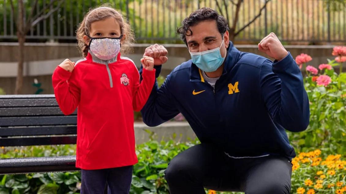 A doctor and a young patient wearing masks pose outdoors, smiling and flexing their arms in a show of strength.
