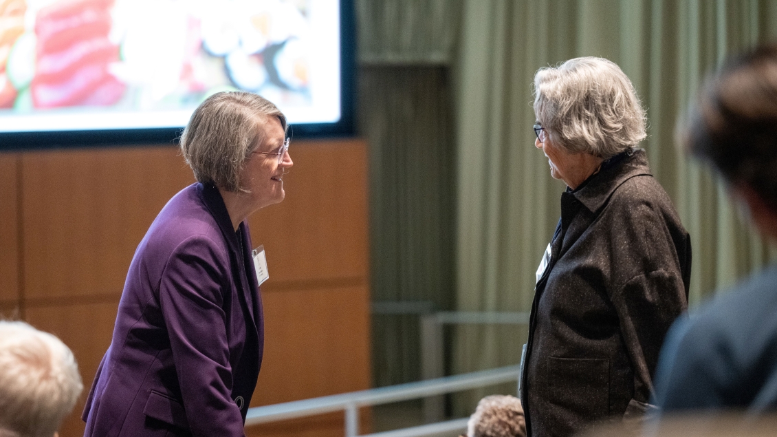 Two people are engaged in conversation in an auditorium with blurred attendees.