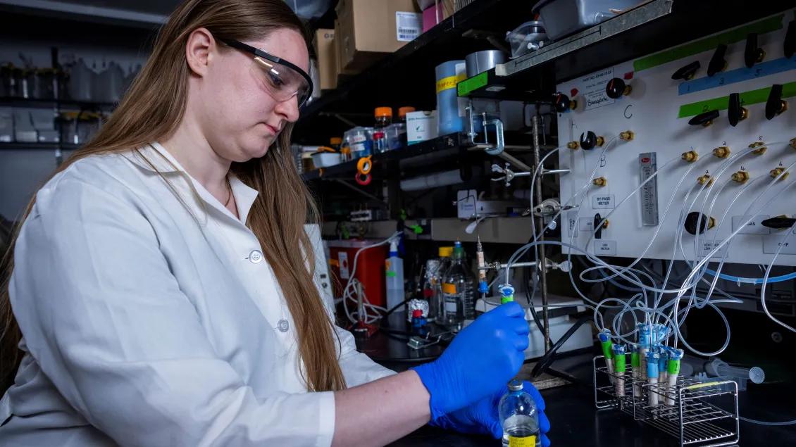 A scientist in a lab coat and safety goggles works attentively at a lab bench, holding a vial.