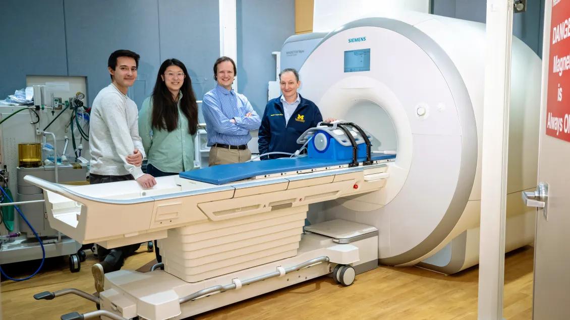 A group of four people stands beside an MRI machine in a medical room.