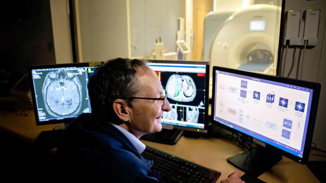Man in a dimly lit room views brain scans on three monitors, with an MRI machine visible behind glass.