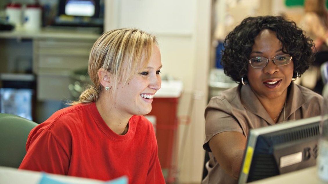 Two colleagues sit side by side at a desk, smiling and looking at a computer screen while discussing information in an office setting.