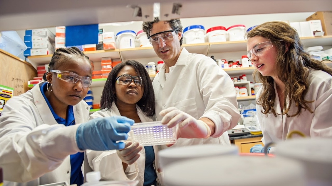 A diverse team of scientists in lab coats and protective eyewear work together at a laboratory bench, examining a sample tray during an experiment.