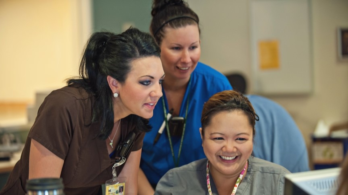 Three healthcare professionals gathered around a computer workstation, smiling and collaborating in a clinical office setting.