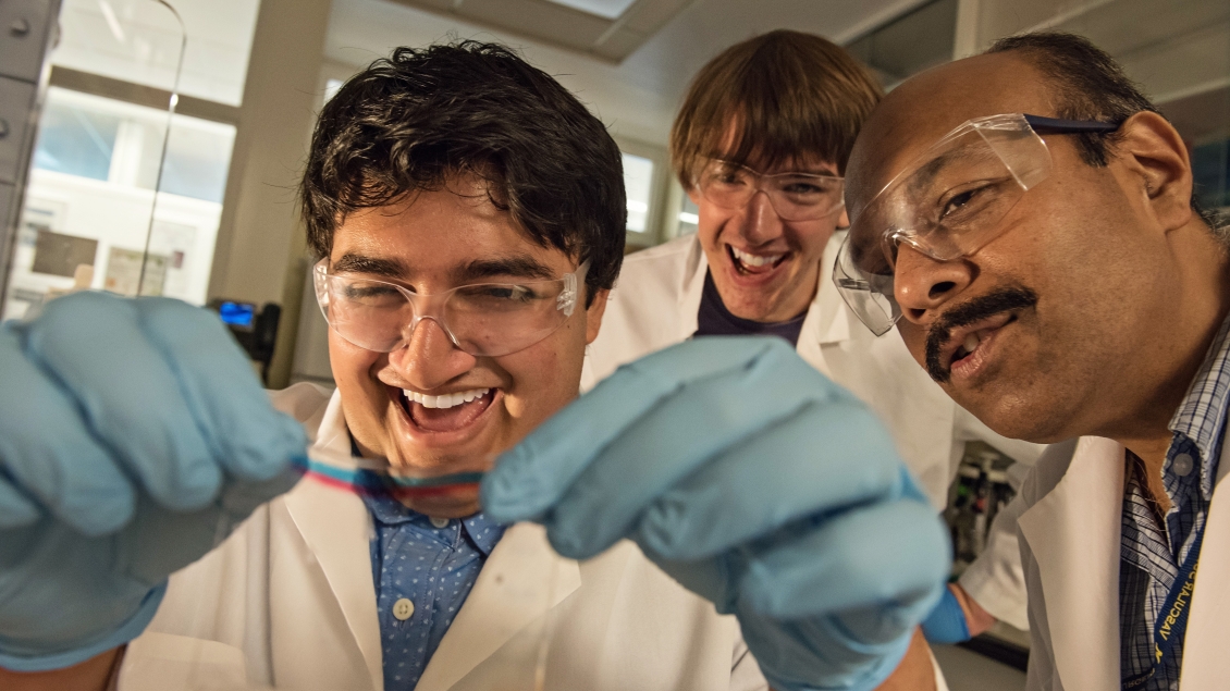 Three researchers wearing lab coats, safety glasses, and gloves smile while closely examining a flexible sample in a laboratory.