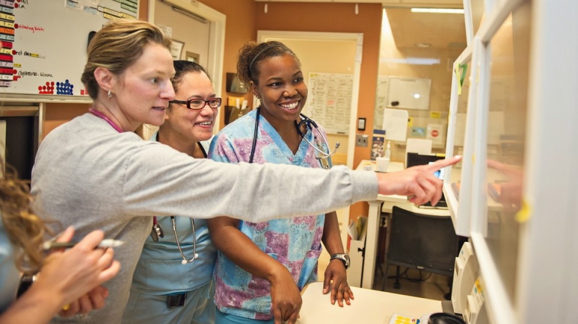 Four healthcare professionals stand together in a clinical workspace, smiling and pointing at a wall-mounted board while reviewing patient or workflow information.