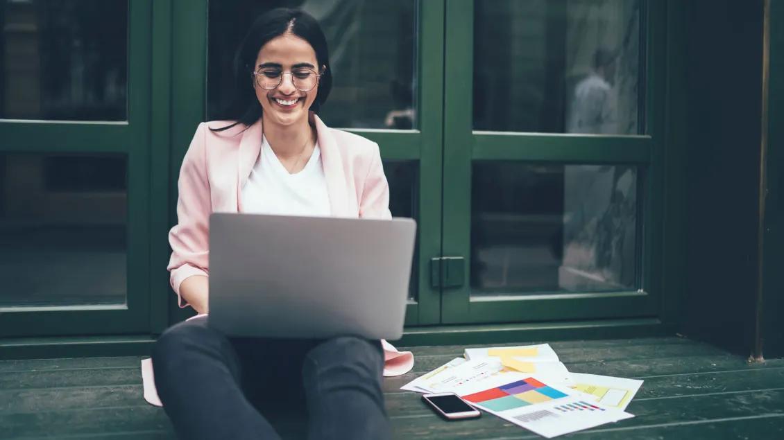 A woman reads publications on her laptop outside a building with her papers & phone beside her