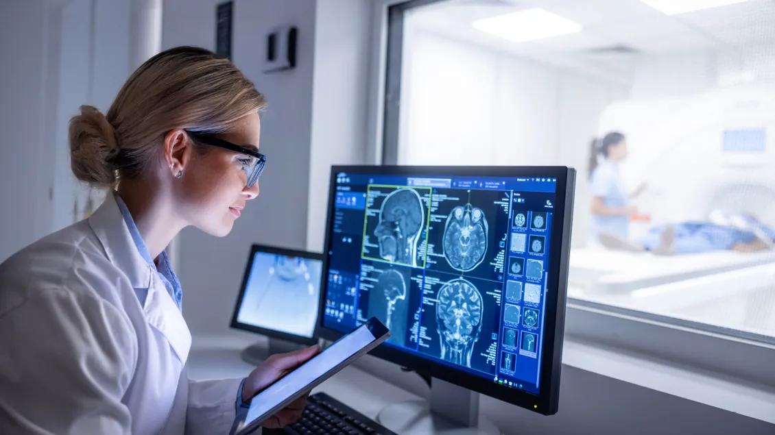 Doctor examining X-ray images on display in MRI control room while in background nurse preparing the patient for examination test.