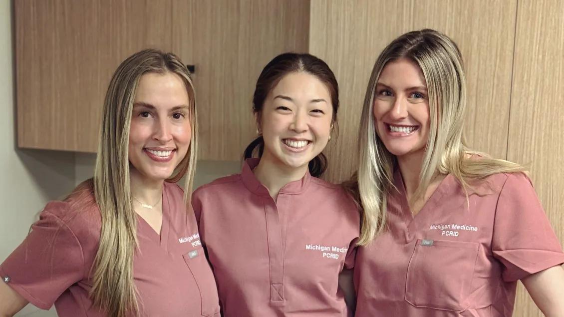 Three smiling healthcare professionals wearing matching pink Michigan Medicine PCRID scrub tops stand side by side in a clinical setting with light wood cabinetry in the background.