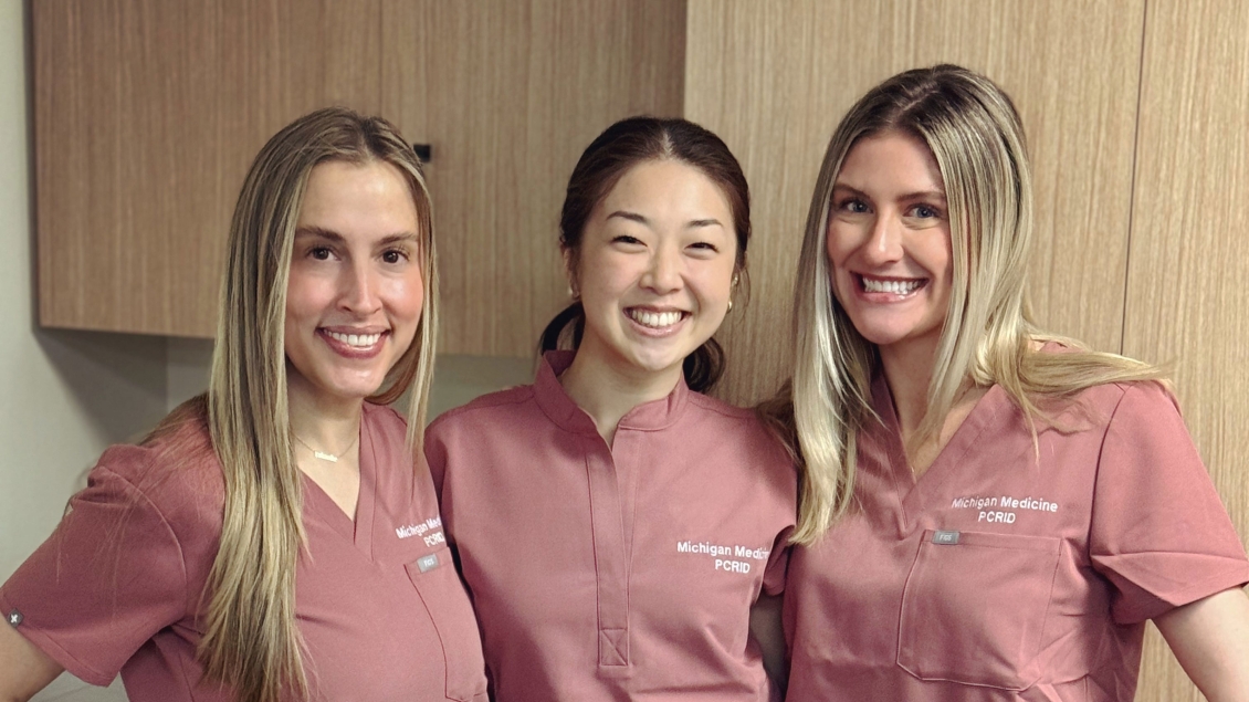 Three smiling healthcare professionals wearing matching pink Michigan Medicine PCRID scrub tops stand side by side in a clinical setting with light wood cabinetry in the background.
