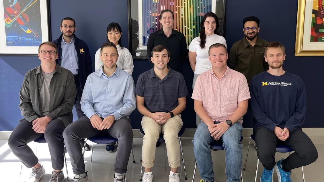 Group portrait of 10 people arranged in two rows, with five seated in front and five standing behind them, in front of a dark blue wall with framed artwork. Several people wear business-casual clothing, and one seated person wears a University of Michigan Medicine sweatshirt. Faces are blurred.