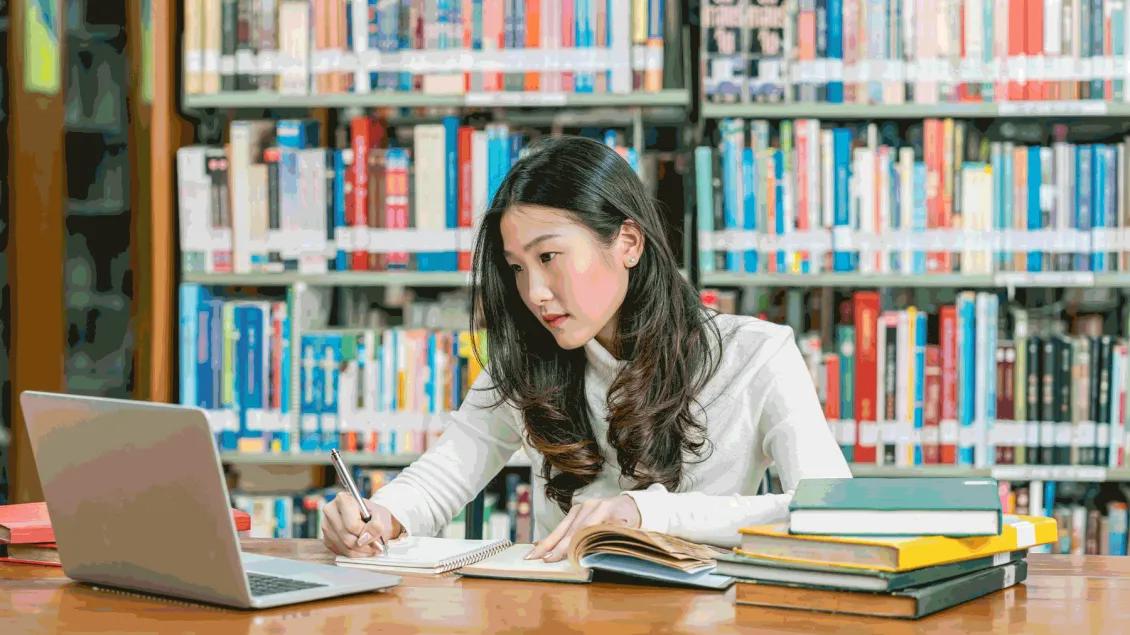 A student reads from a computer while taking notes at a library