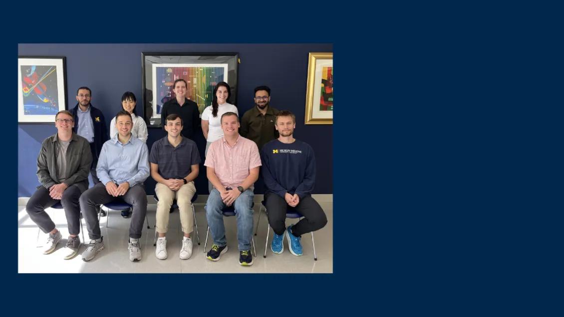 Group photo of nine people posing in front of a dark blue wall with framed artwork.