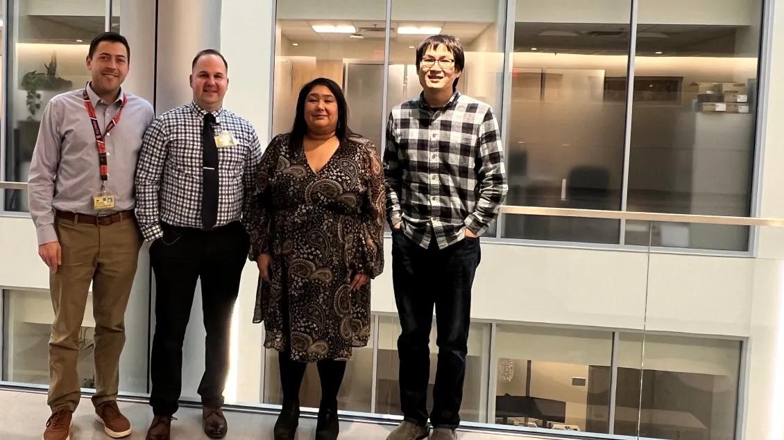 Four people standing side-by-side in front of tall glass windows with office spaces behind the glass