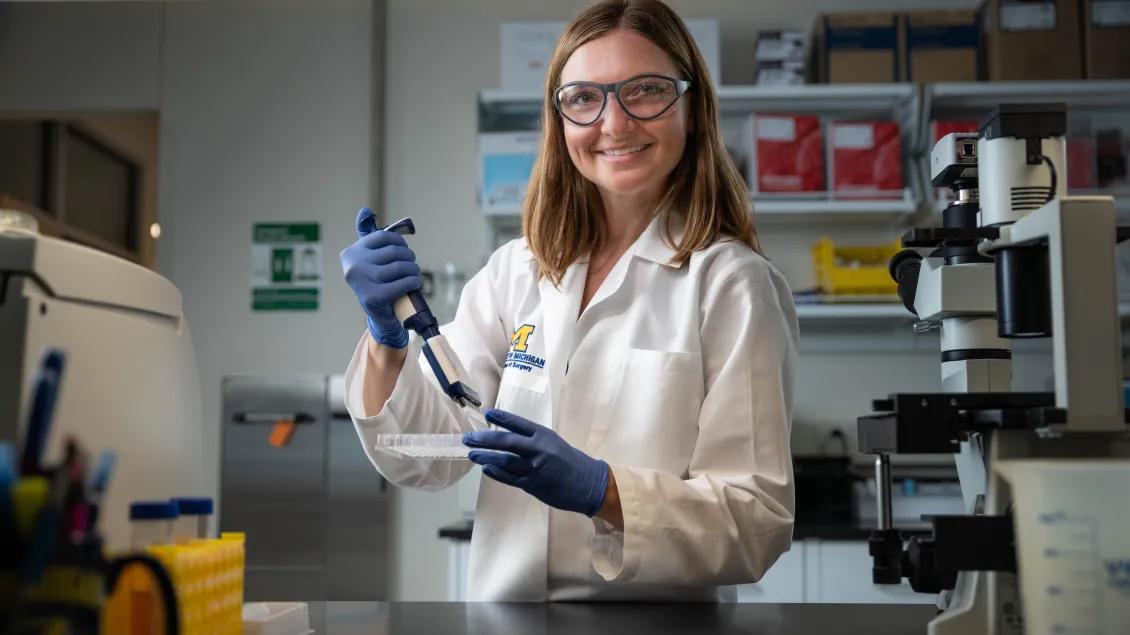 A person in a white lab coat smiles while using a pipette in a lab.