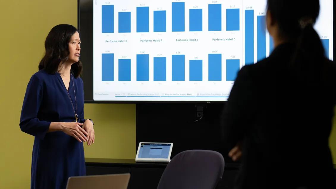A person stands beside a large screen displaying blue bar graphs as another person looks on in the foreground.