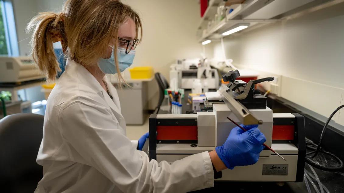Researcher in a lab coat, mask, and gloves operates laboratory equipment