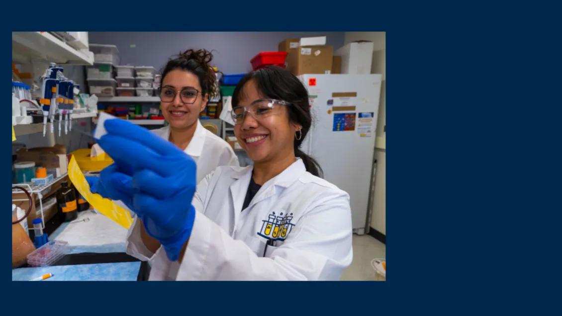 Two people smiling while wearing white coats and working in a research lab